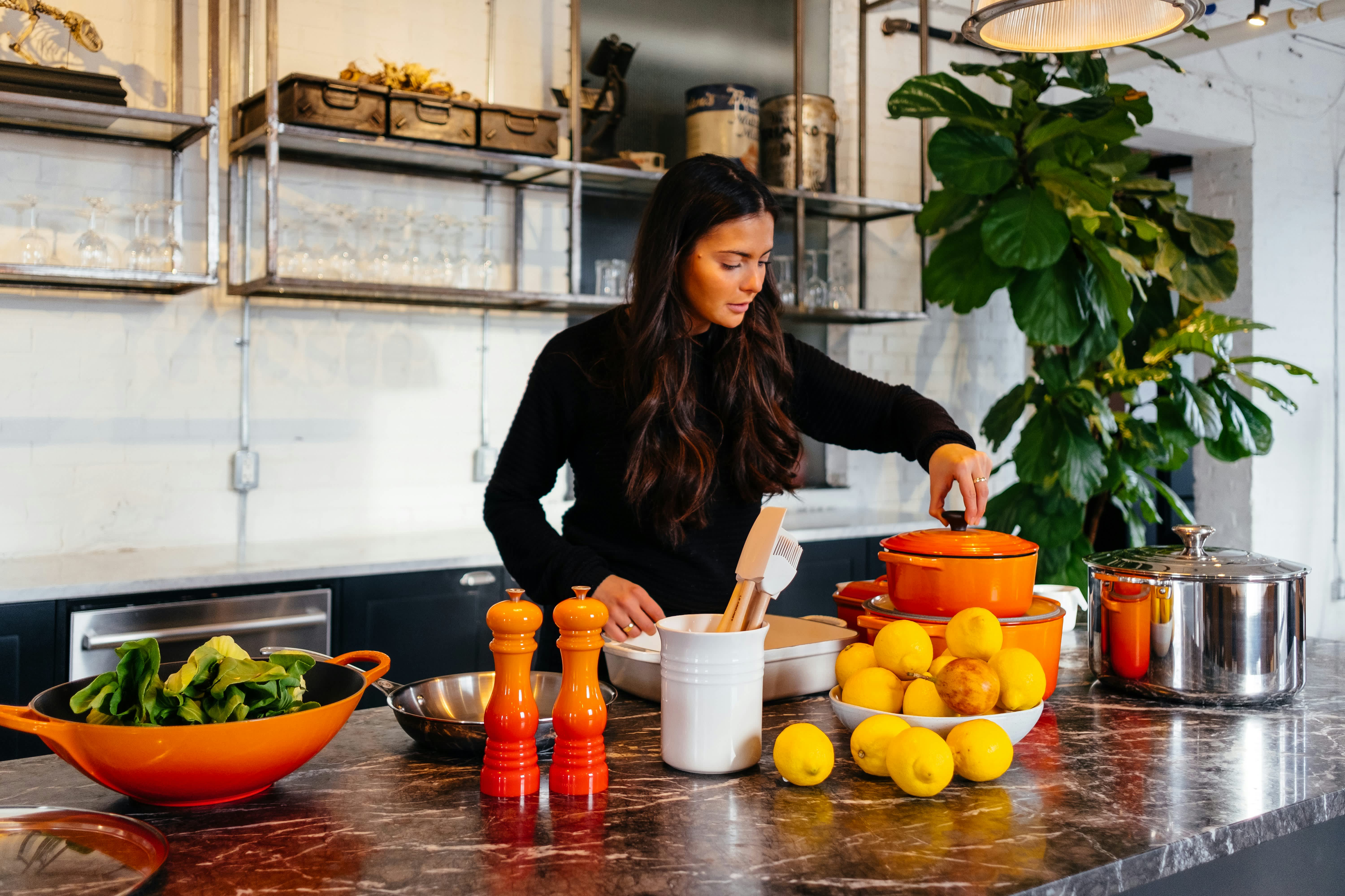 Woman preparing food in a modern kitchen surrounded by orange cookware, lemons, and fresh vegetables.