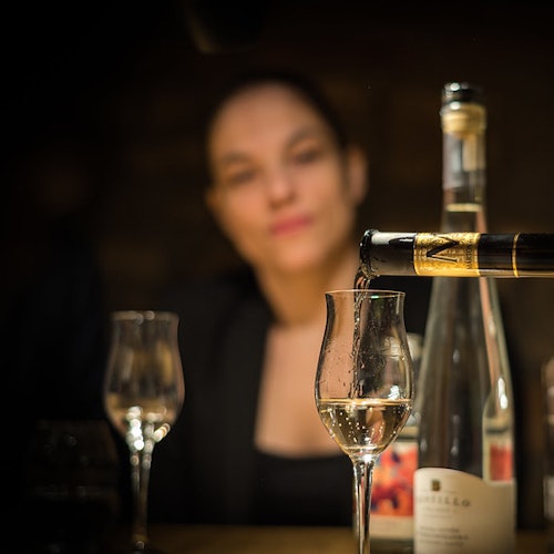 Una persona sullo sfondo, con il focus su un bicchiere che viene riempito di vino bianco da una bottiglia. Altri due bicchieri e bottiglie nelle vicinanze.
