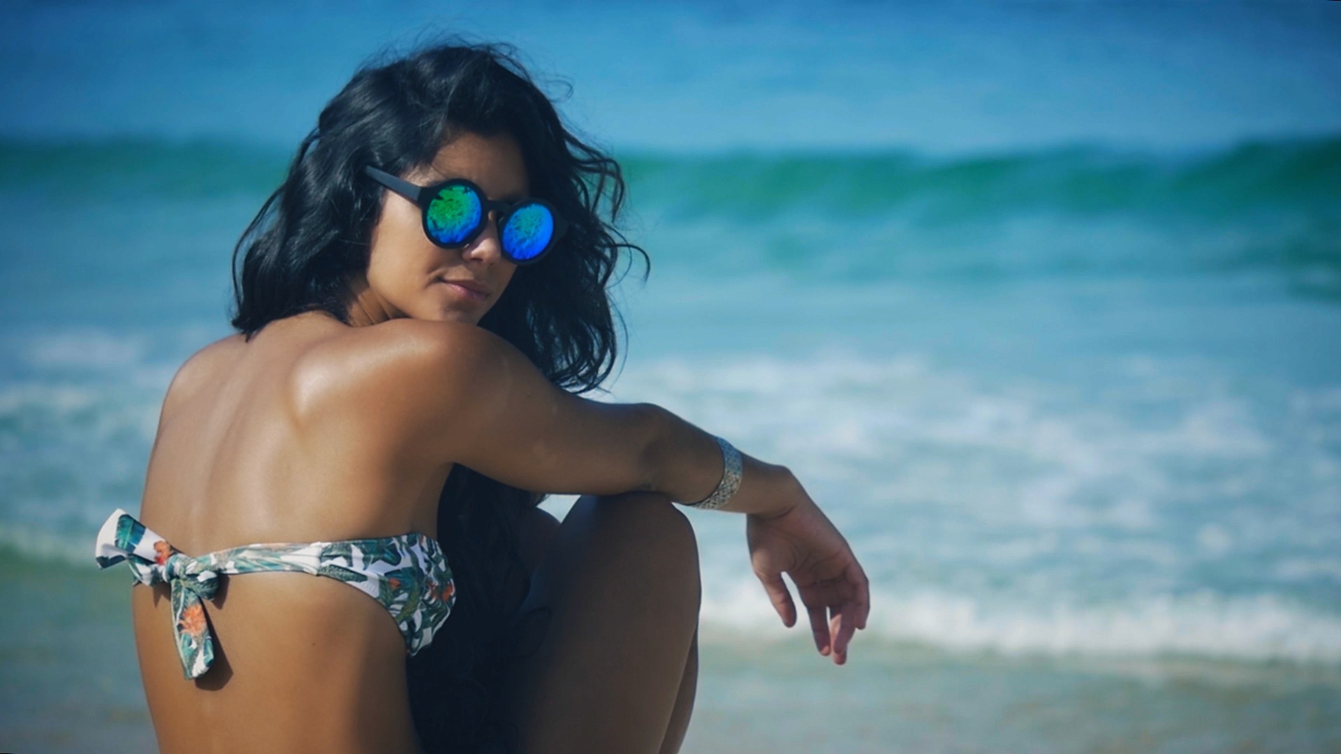 Editorial fashion film of a woman looking at the camera with sunglasses wearing a bikini at the beach