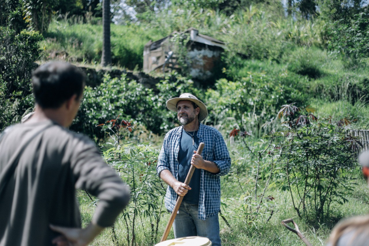 Homem de chapéu segurando uma ferramenta conversa com participantes em uma horta, com casa de madeira ao fundo