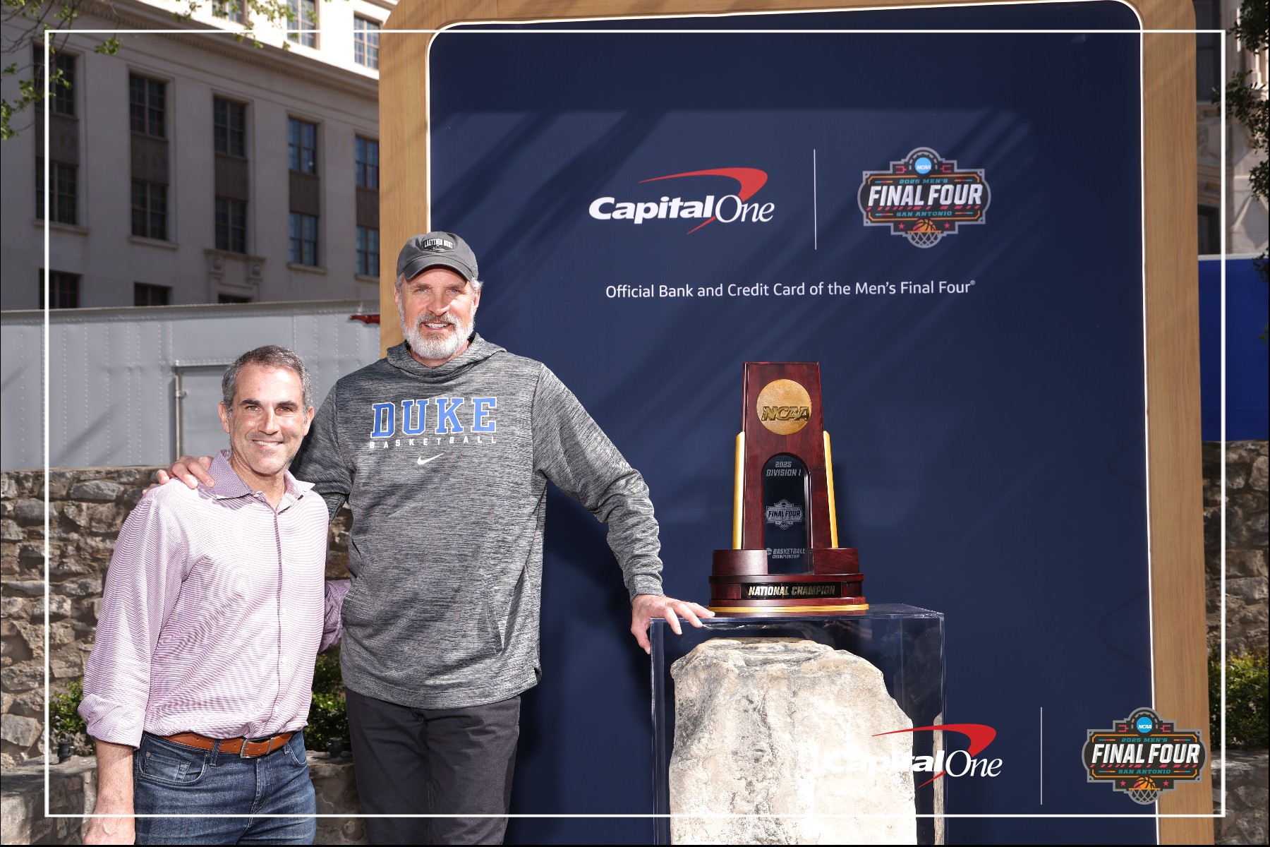 Christian Laettner stand next to a NCAA Final Four trophy display in front of a branded Capital One and Final Four backdrop outdoors. One man wears a Duke sweatshirt and cap; both are smiling at the camera.