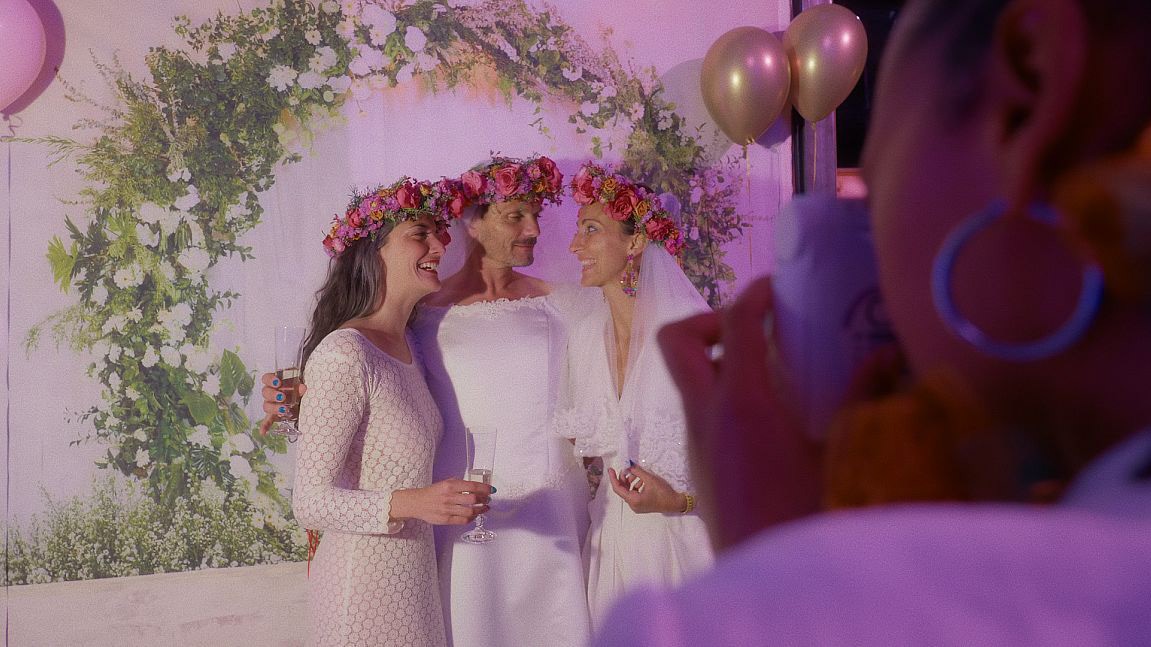 Three people stand together, smiling in front of a floral backdrop, surrounded by soft purple lighting.
