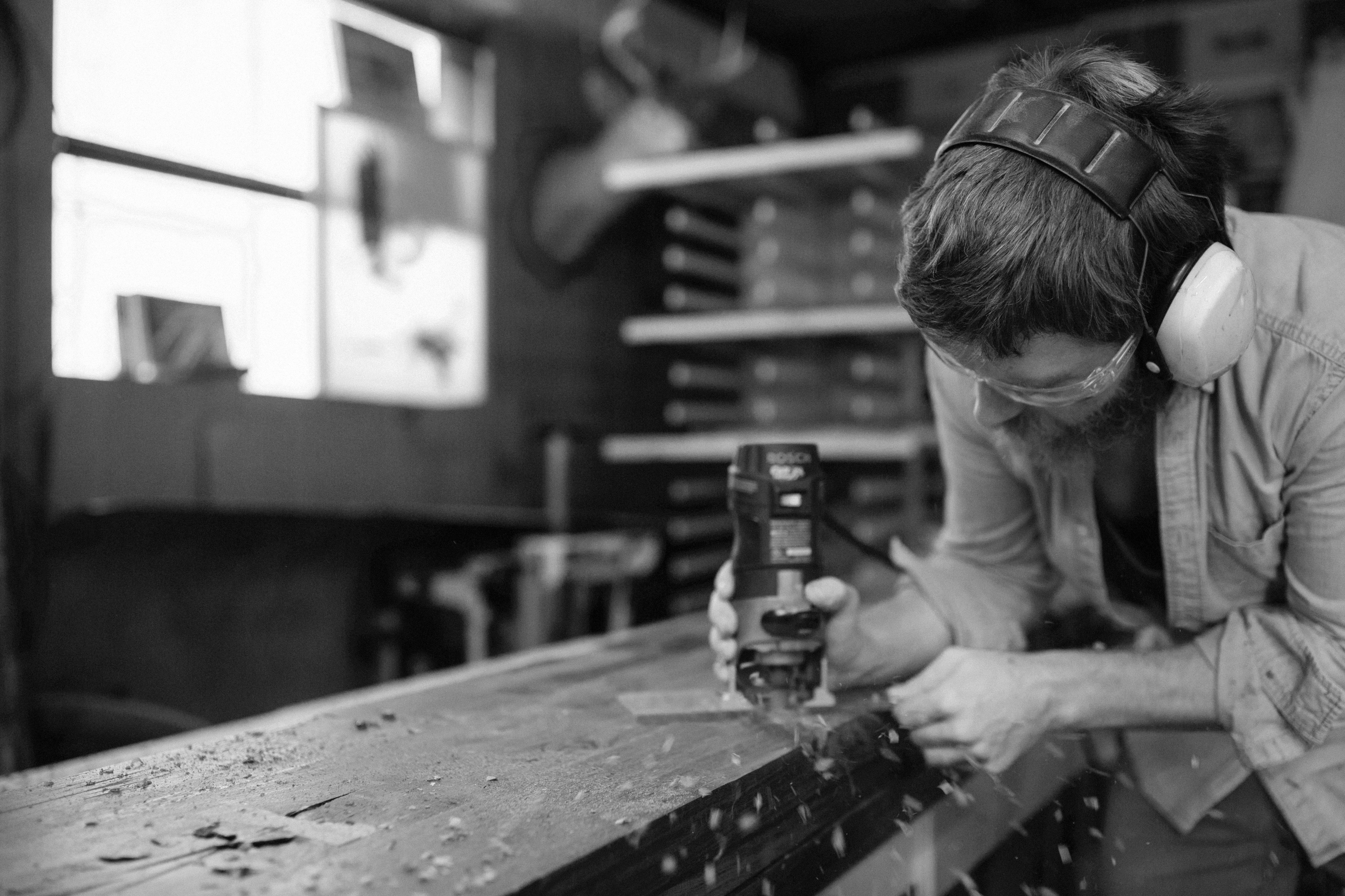 Ein Schreiner bearbeitet ein massives Holzwerkstück mit einer Oberfräse in seiner Werkstatt, Holzspäne fliegen, dokumentarische Schwarz-Weiß-Fotografie.