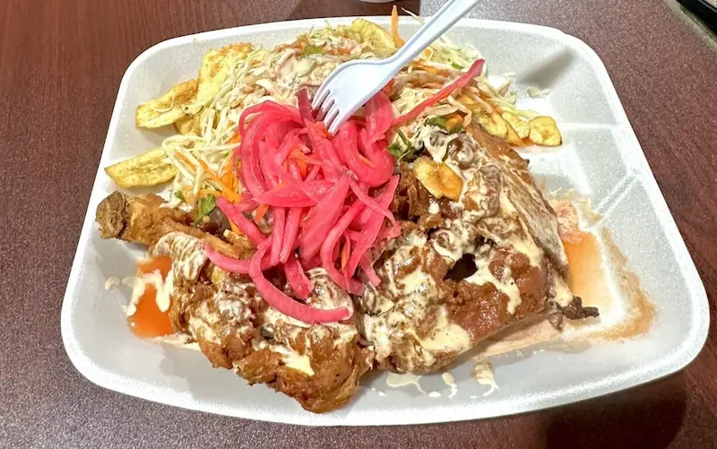 A styrofoam plate filled with crispy fried chicken topped with pickled red onions, alongside seasoned fries and a fresh cabbage slaw, sits on a wooden table, highlighting a mouthwatering meal available at the Oneco Farmers Market in Bradenton, FL.