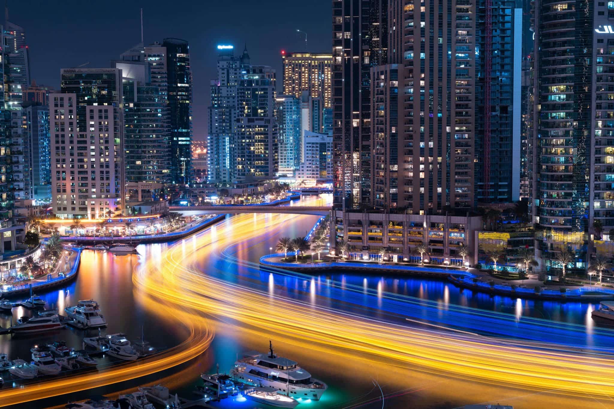 Night view of Dubai Marina with illuminated skyscrapers, bright light trails on the water, and boats docked along the marina.