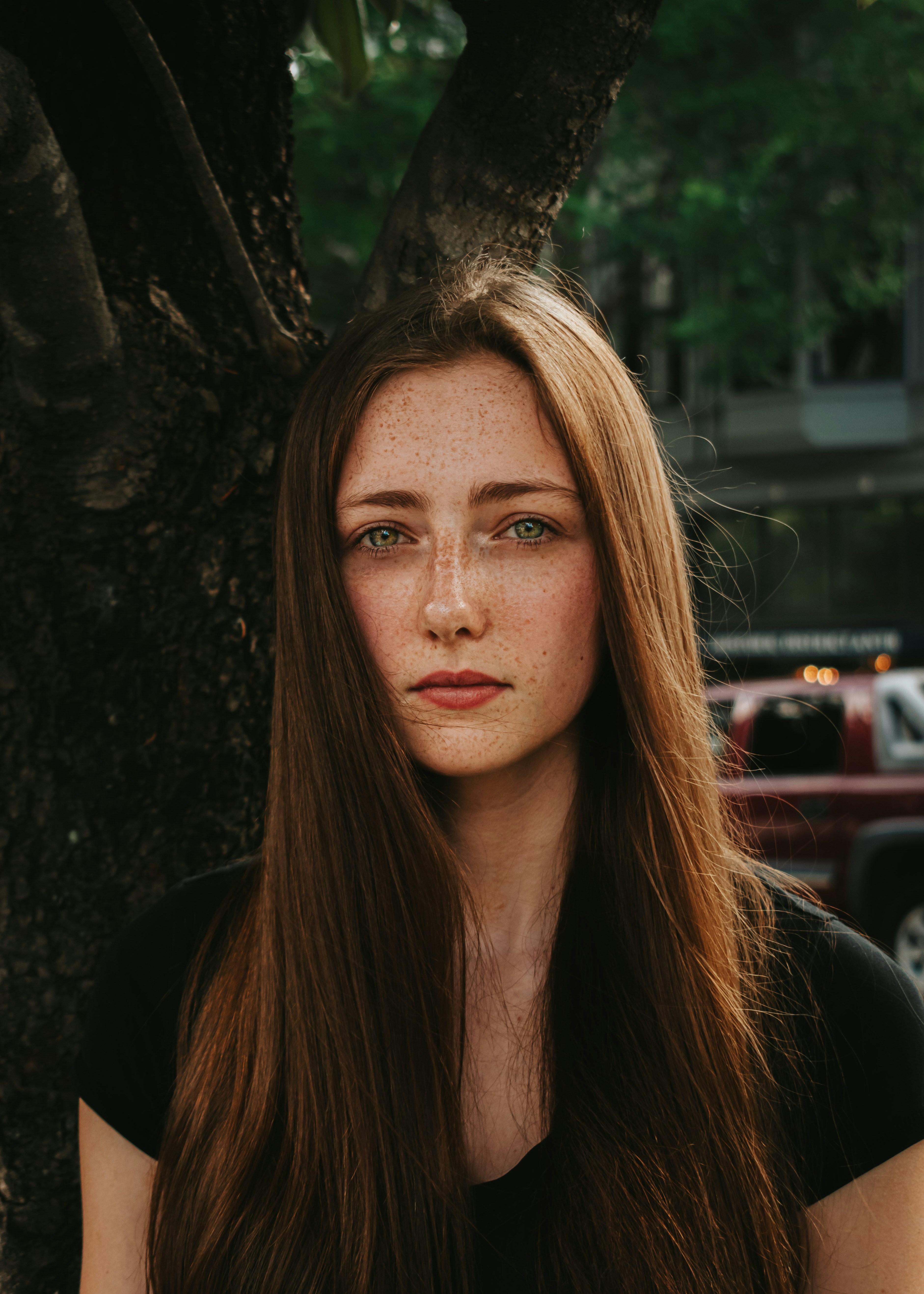 A young woman with long hair stands outdoors, looking directly at the camera with a neutral expression.
