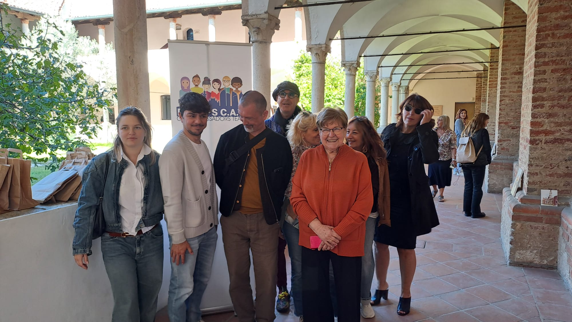 A group of people is gathered in a covered walkway with arches, standing in front of a banner on a sunny day.
