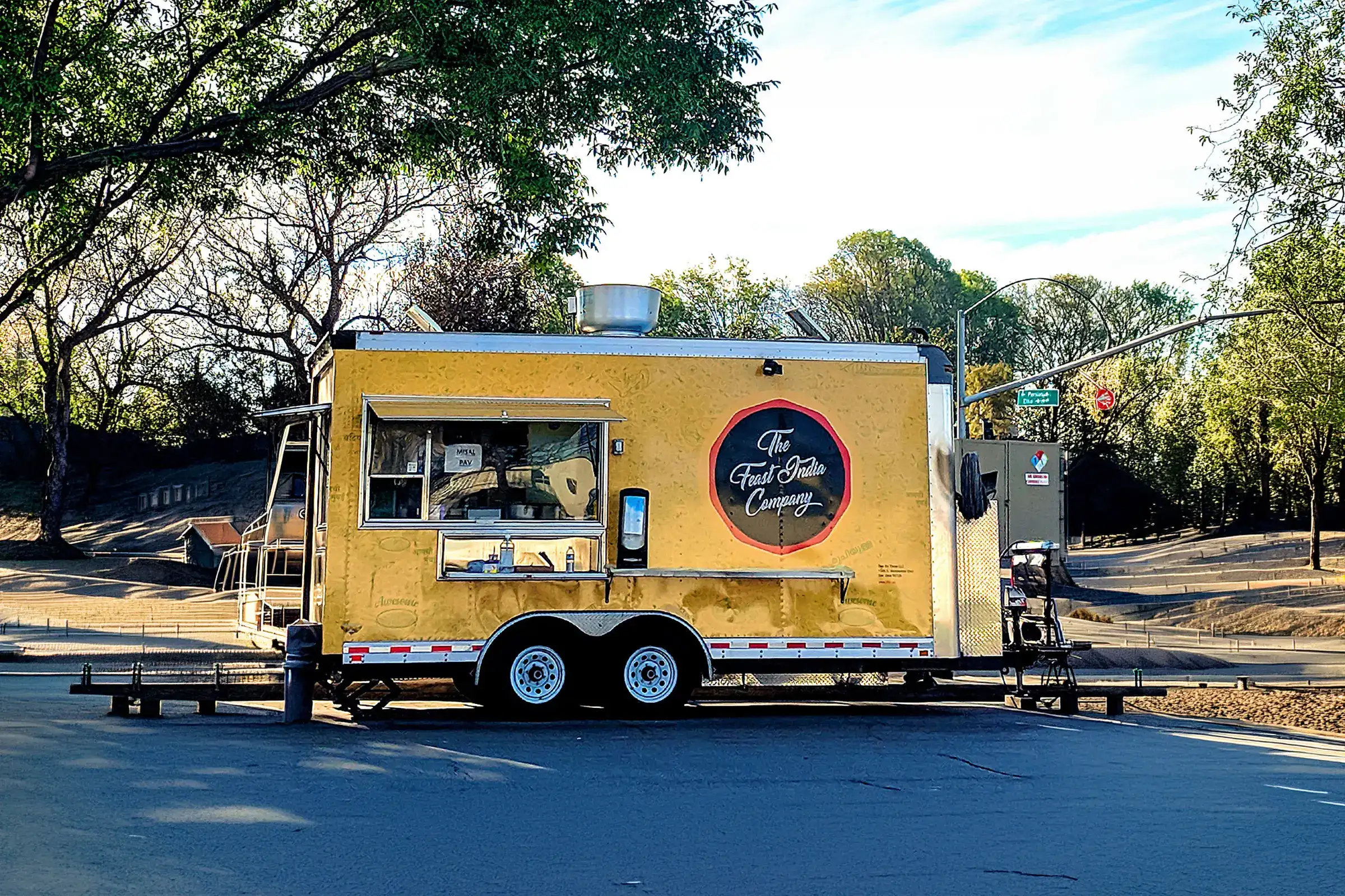 Yellow food truck with a serving window and “The Happy Camper” logo parked on a sunny, tree-lined street near an intersection.