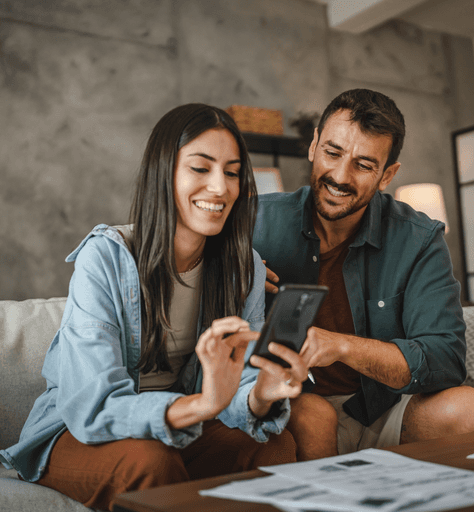 Casal sentado em um sofá sorrindo para o celular após contratar um seguro para smartphone.