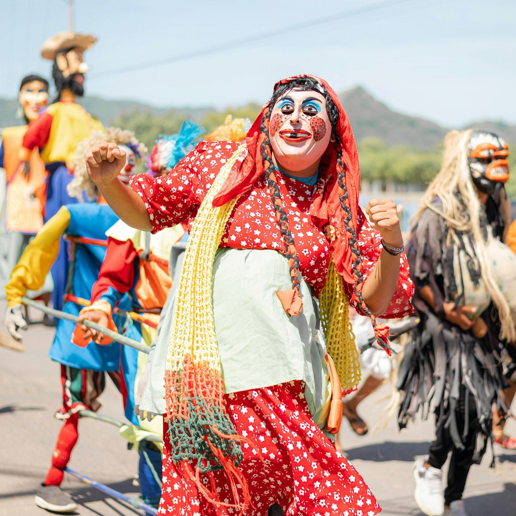 A vibrant group of people dressed in colorful costumes and masks participate in a lively cultural parade, showcasing traditional attire and joyous celebrations in an outdoor setting.