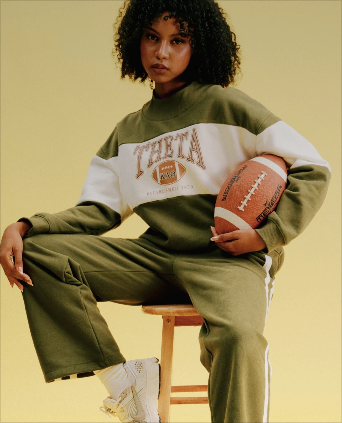 Model holding a football while wearing an olive-and-cream Theta sweatshirt and matching pants.