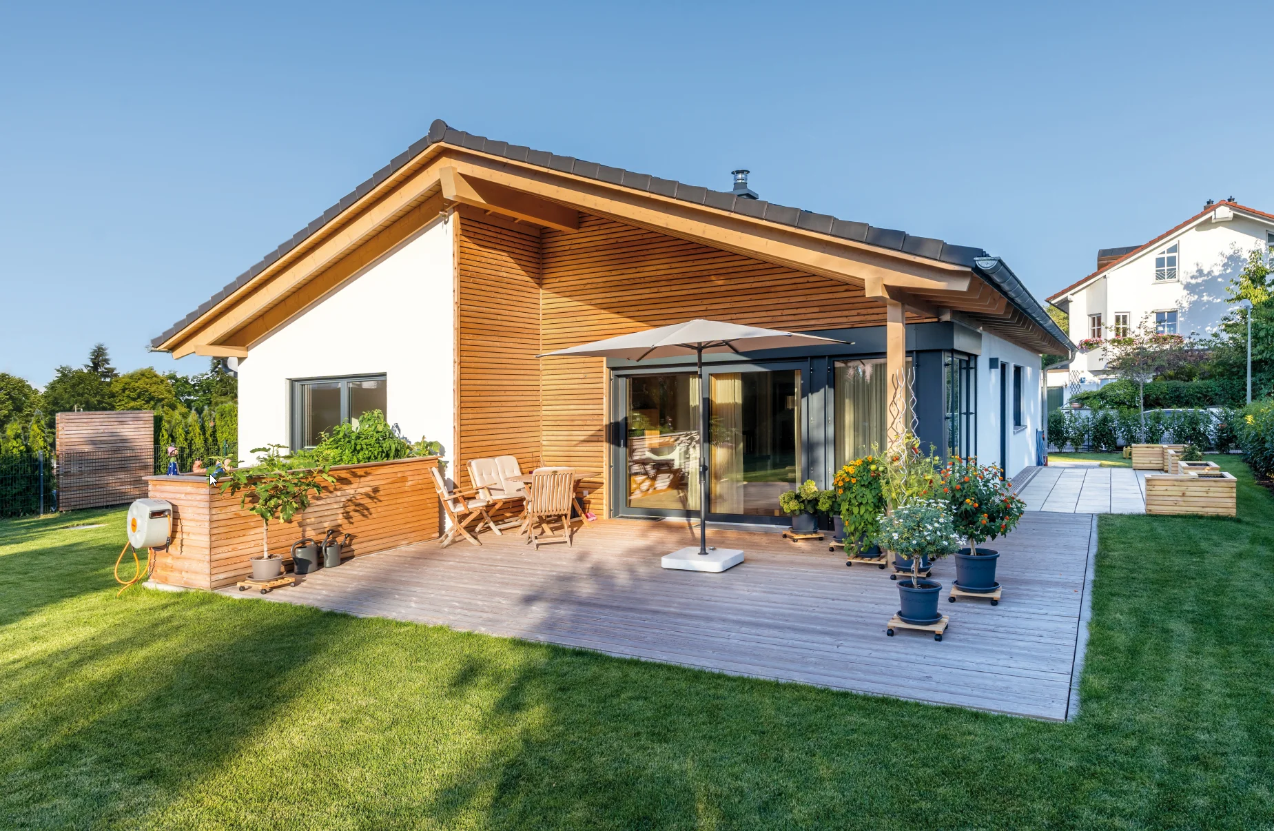 Modernes Einfamilienhaus mit Holzfassade, überdachter Terrasse mit Möbeln sowie Garten mit Rasen und Blumenkübeln.