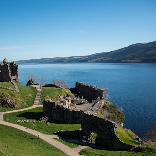 在湛蓝天空下，石砌城堡的废墟俯瞰着一片广阔而宁静的湖泊，背景是连绵起伏的山丘。