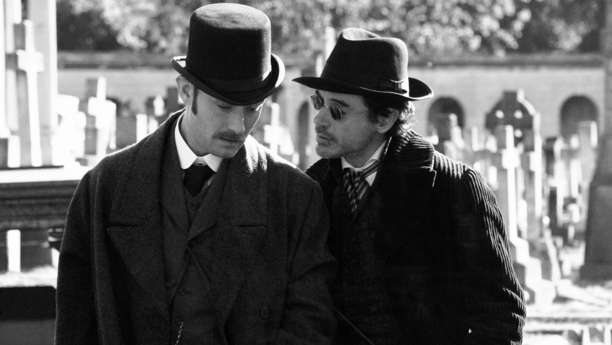 black and white photo of 2 victorian gentlemans dressed elegantly for winter. One of them is whispering to the other's while walking on a cemetery alley