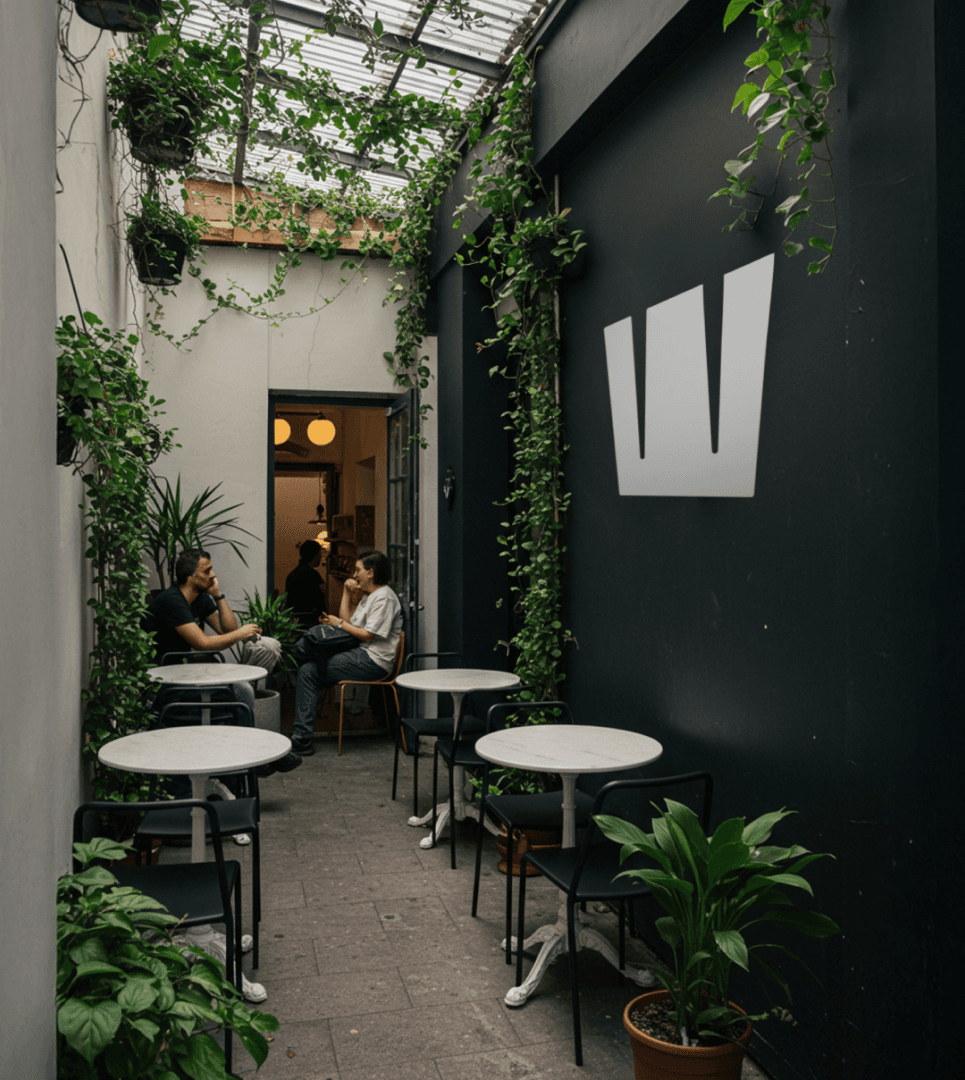 Cozy alleyway café with small round tables and chairs, surrounded by lush green plants under a glass roof, and a simple geometric wall design.