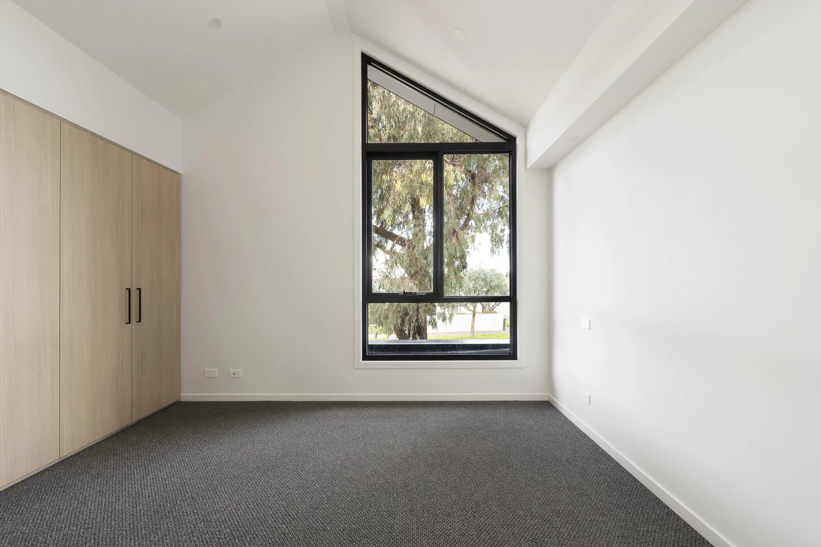Bedroom with pale-oak built-in wardrobes, grey carpet and a raked ceiling with an angled black-framed window framing a gum tree and garden