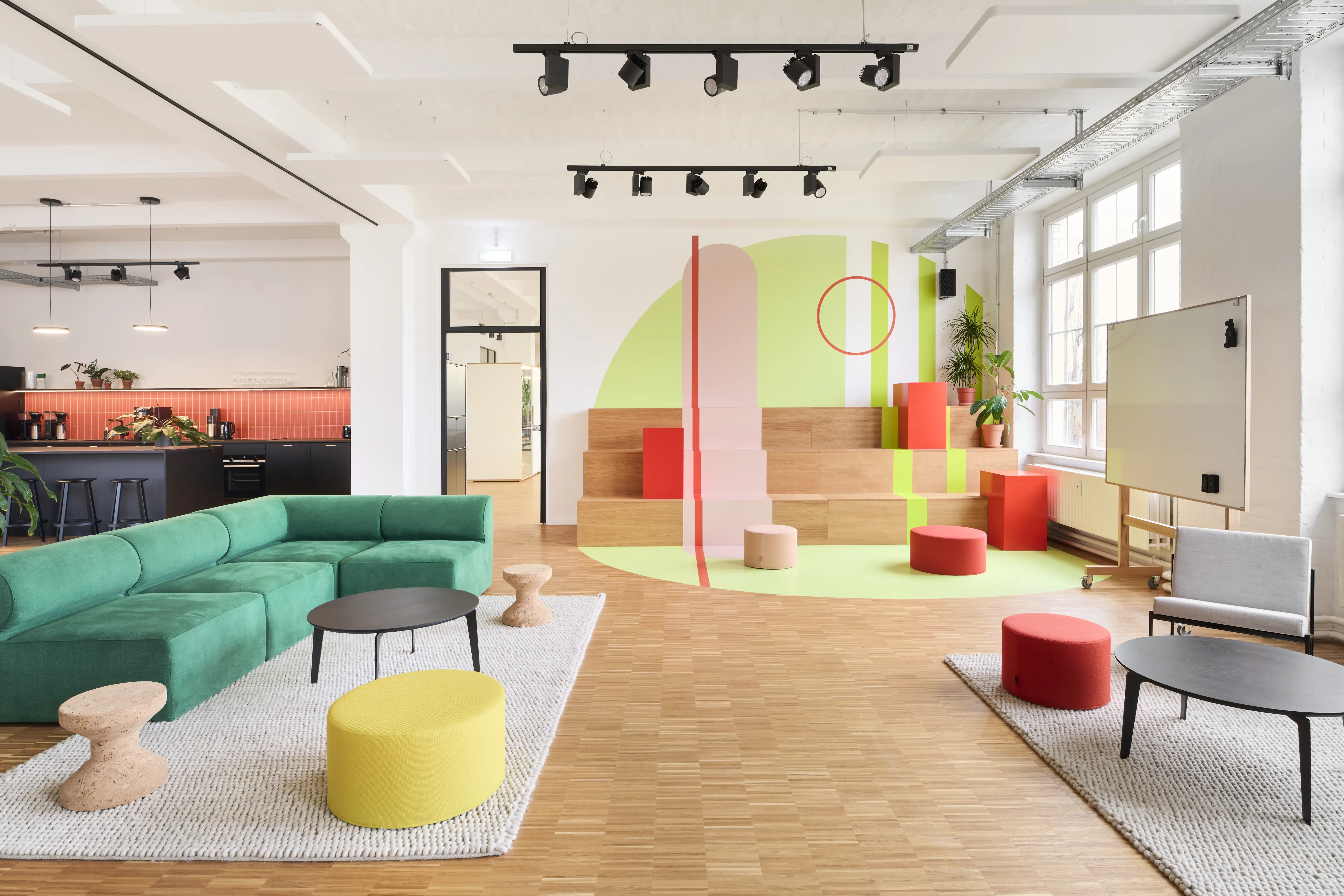 Lounge and play area at PLAN A with a green corner sofa, cork stools, yellow and red poufs, an oak seating tribune with abstract wall mural in green, pink and red, and a mobile whiteboard.