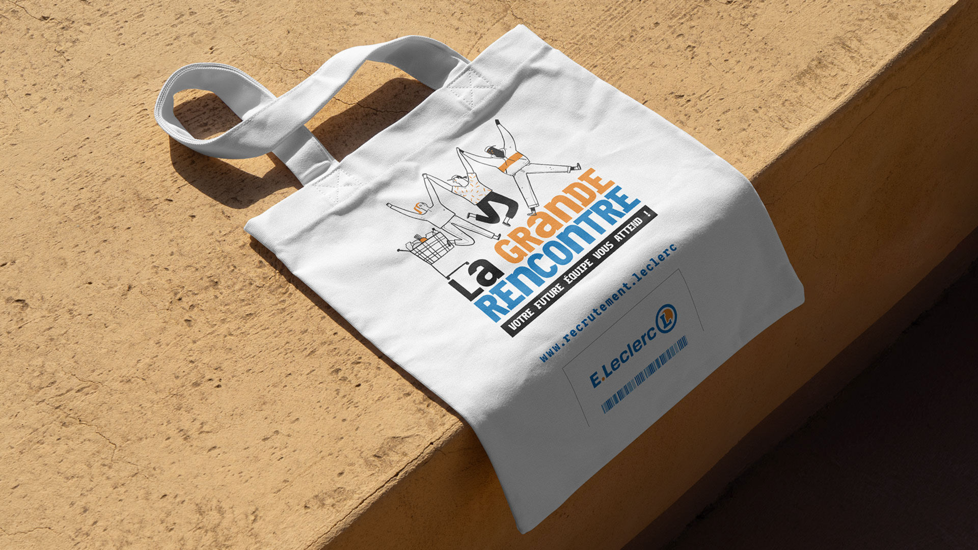 Tote bag en tissu blanc de l'enseigne E.Leclerc pour une opération de recrutement appelée "La Grande Rencontre", posé sur une surface en béton clair.