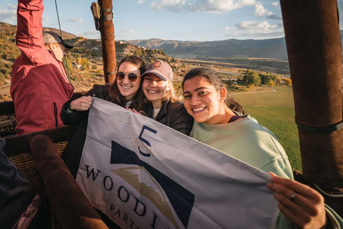 Three retreat guests hold a Woodline Partners anniversary banner inside a hot air balloon basket.