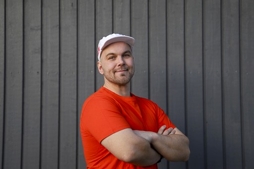 Yrittäjä Joni Salmi tummanharmaan lautaseinän edessä, oranssi t-paita ja valkoinen maalarinlakki, hymyilevä ja itsevarma asenne.