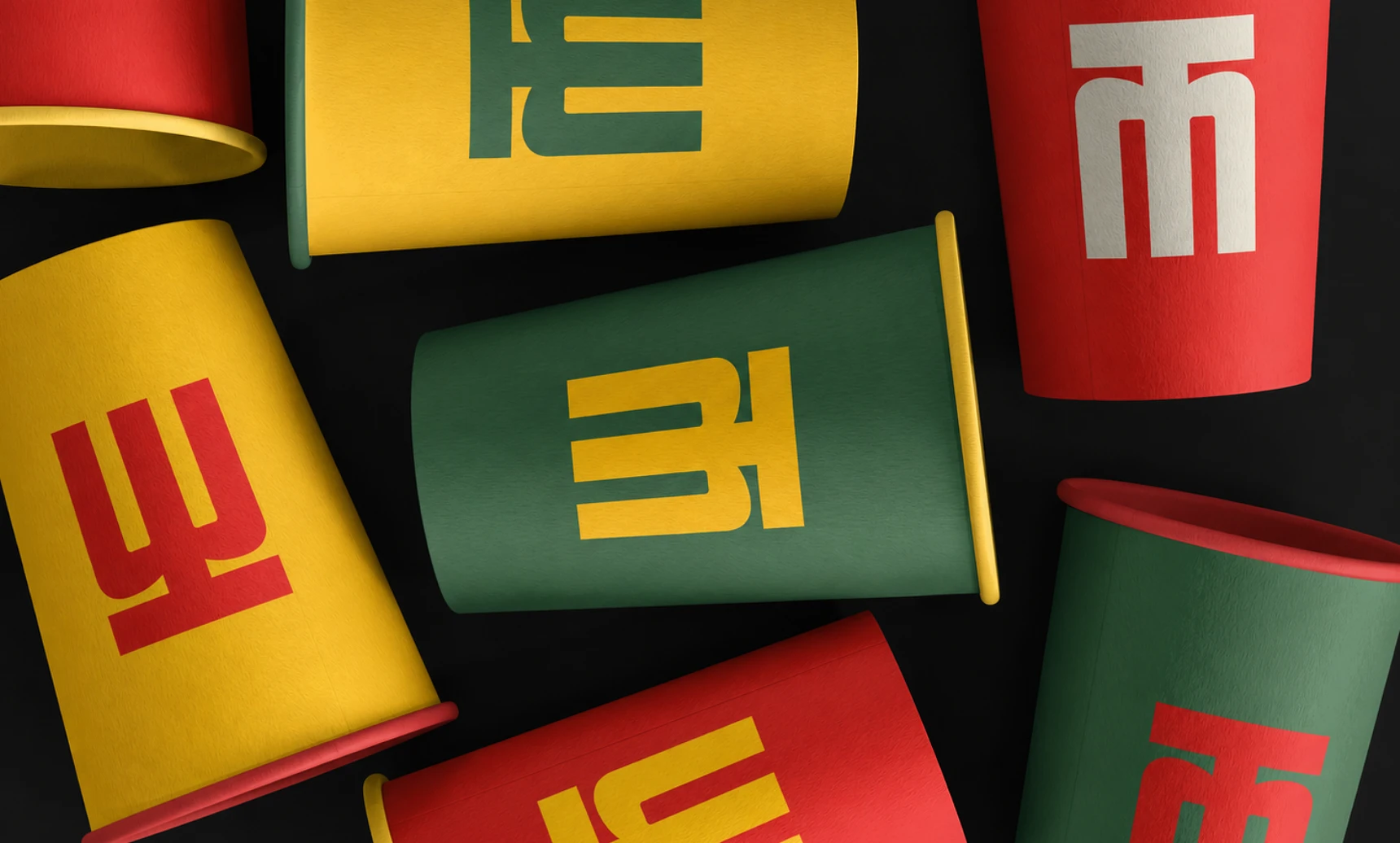 Scattered red, yellow and green paper cups on black, each printed with part of the Mangaluru Canteen monogram.