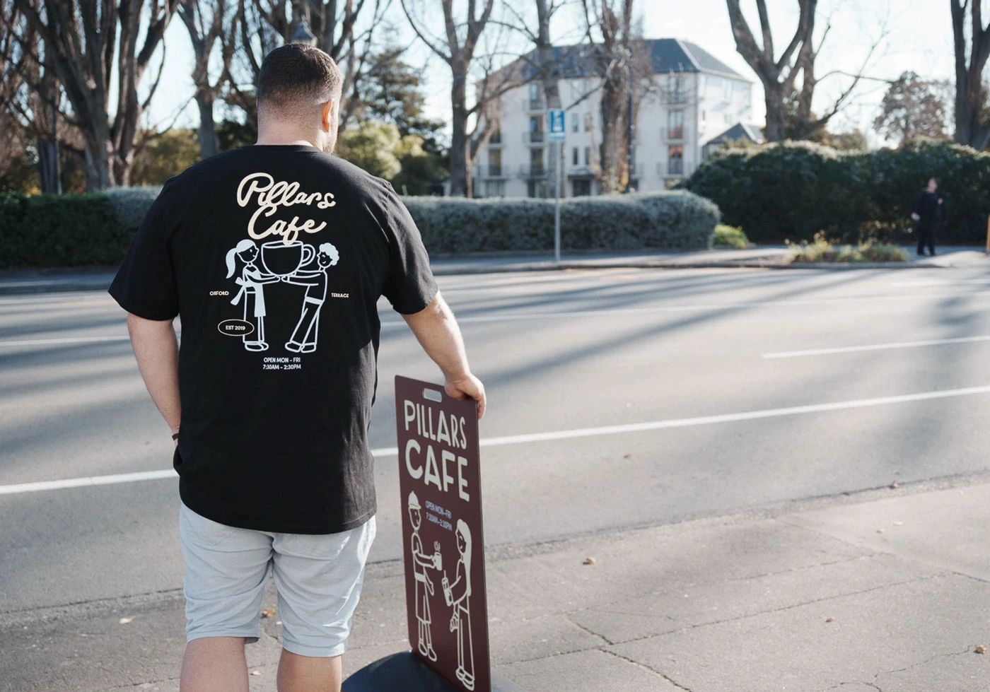Person carrying a Pillars Cafe sandwich-board sign while wearing a branded black T-shirt.
