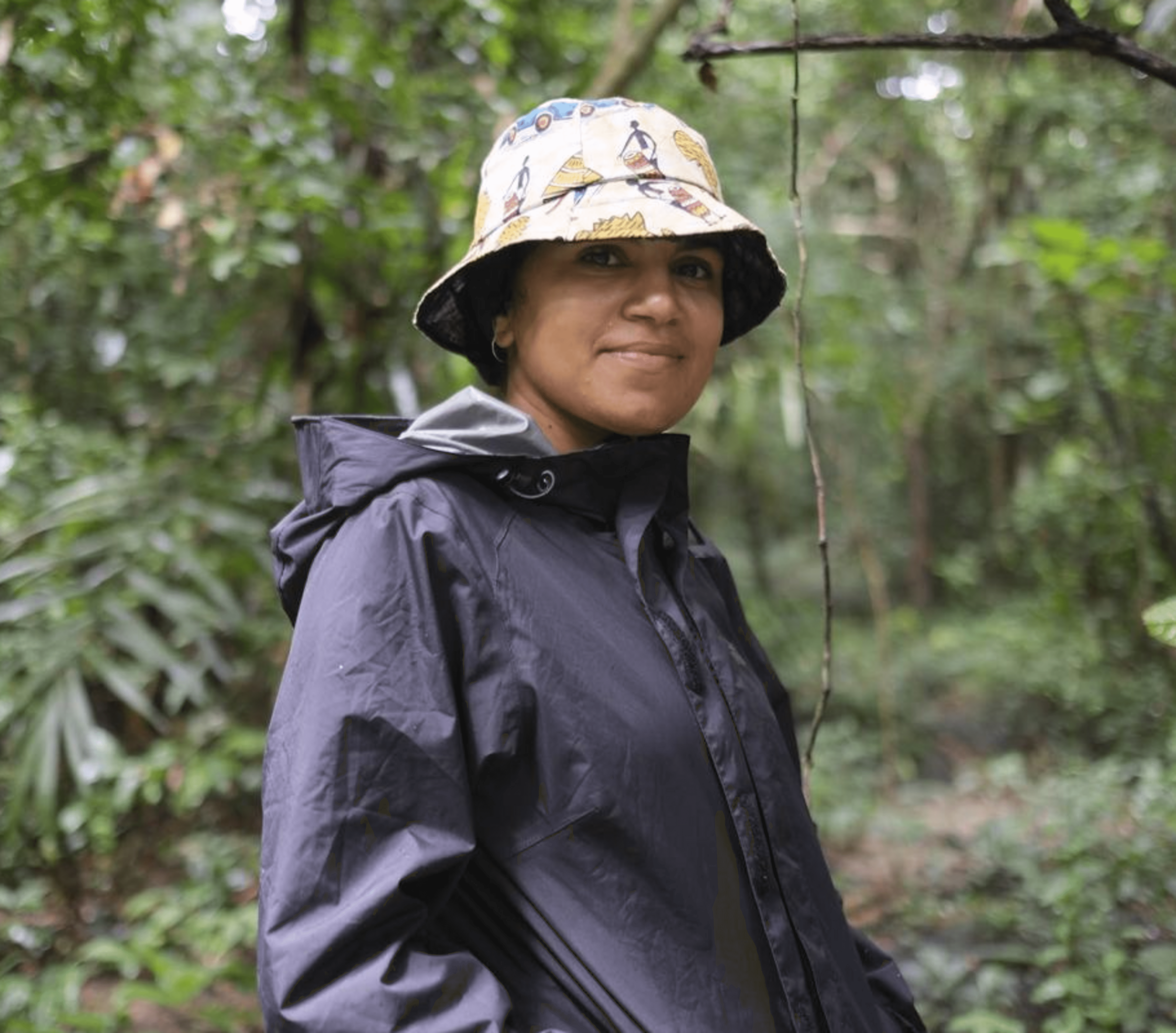 A woman with a soft smile, wearing a hat and rain jacket, standing in a rainforest.