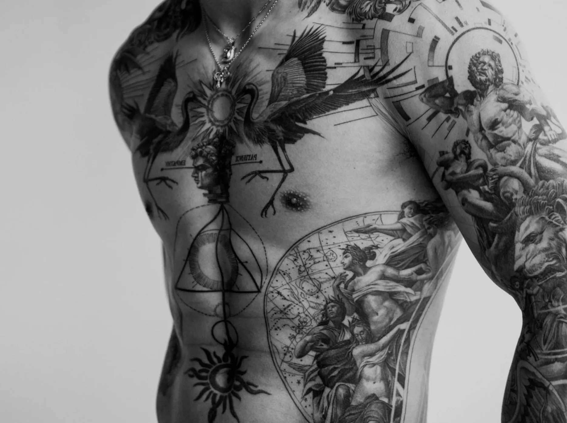 A man with a complex chest tattoo that includes mythological figures, celestial bodies, and geometric patterns.