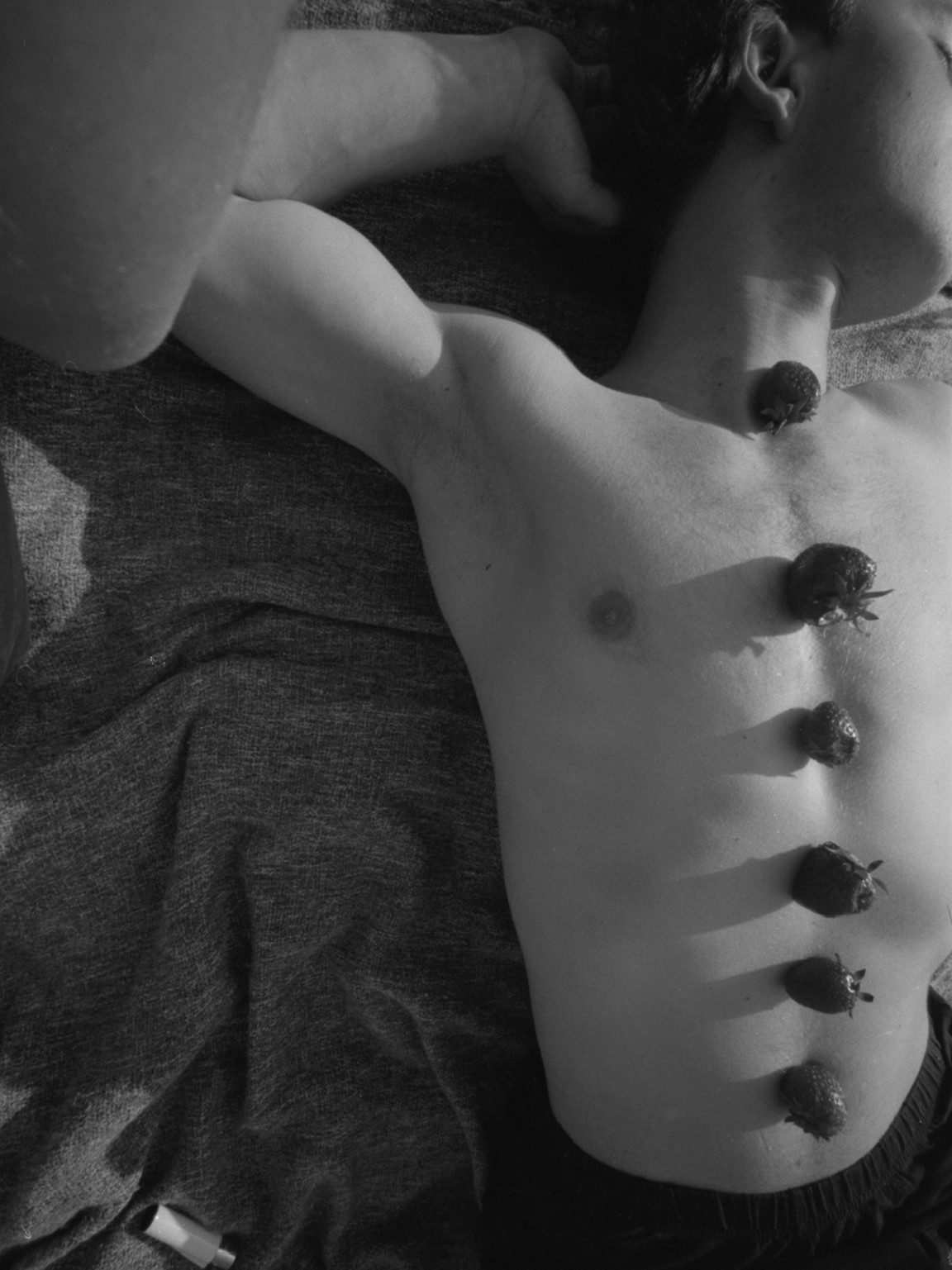 "Man lying on his back with arms raised, strawberries arranged from neck to navel along his torso"