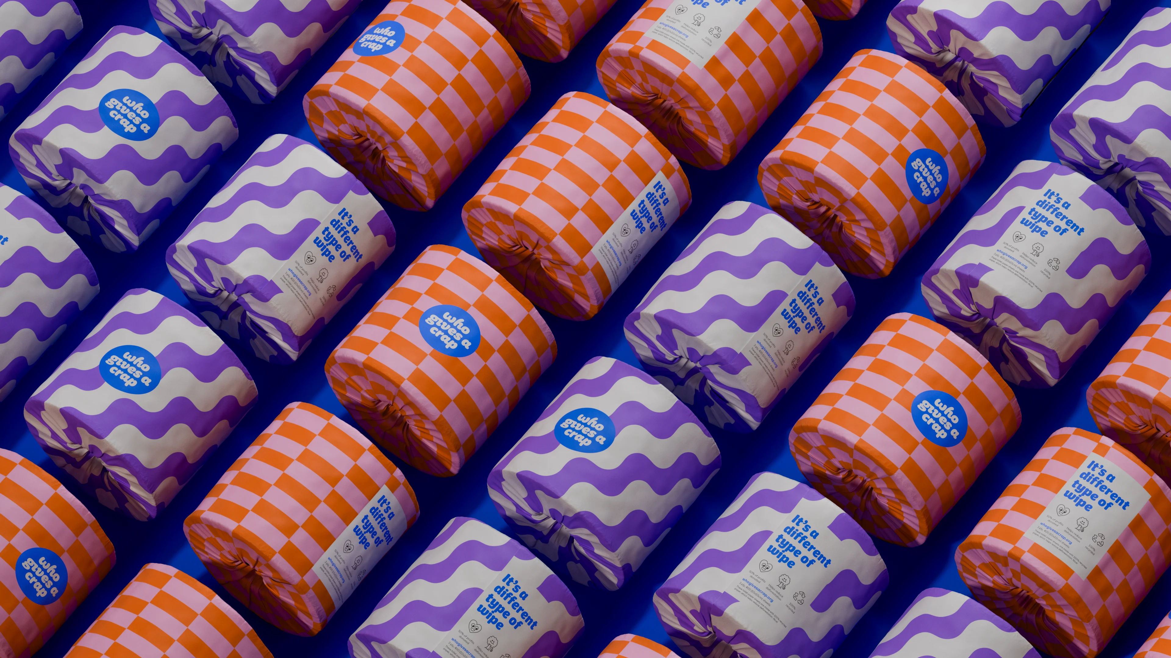 Diagonal rows of Who Gives A Crap rolls in purple wave and orange check wrappers on blue, reading It's a different type of wipe