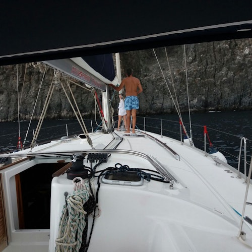 Two people stand on the deck of a sailboat near rocky cliffs. The one closer to the camera is shirtless and wearing blue shorts.