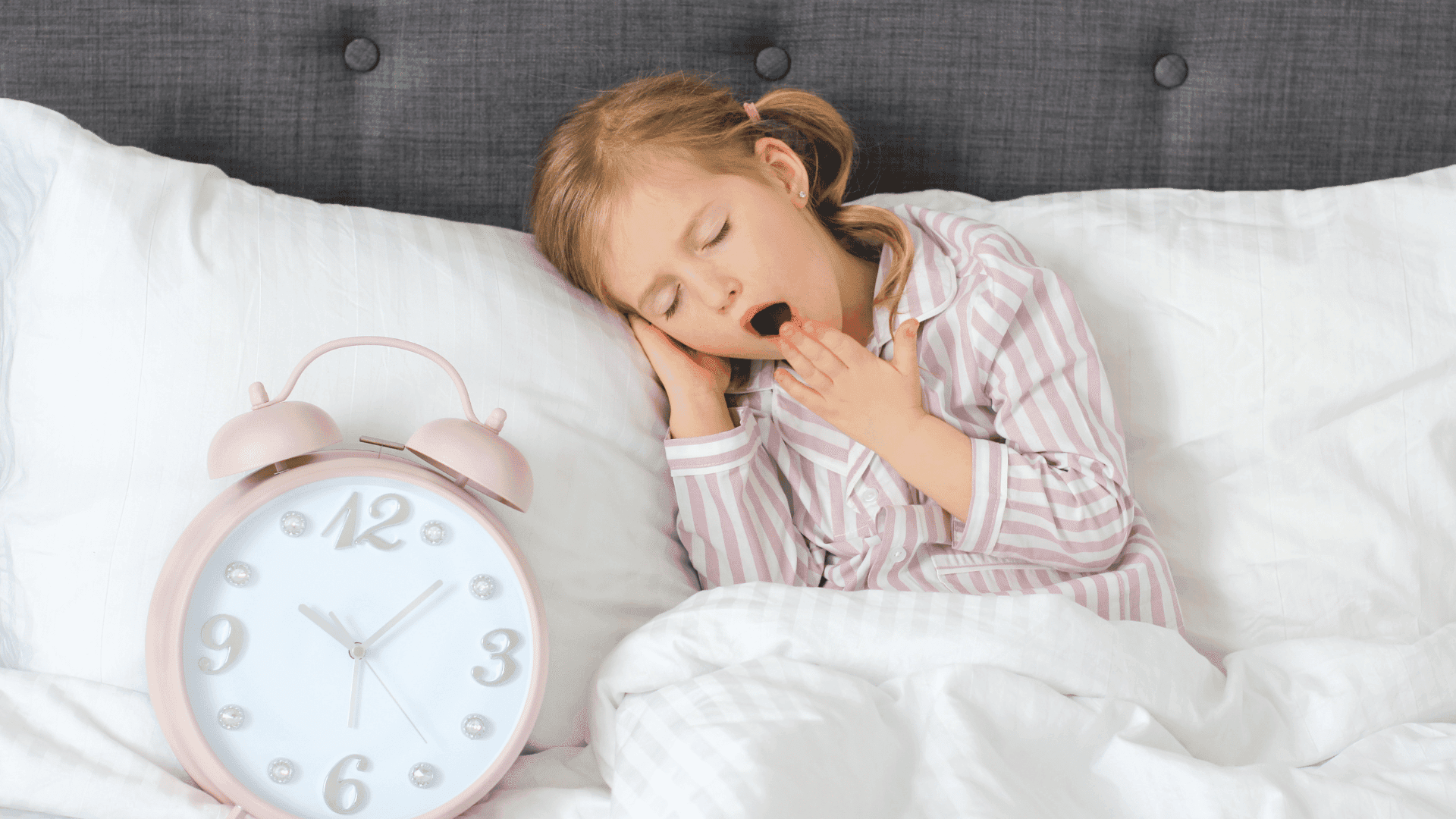 A young girl in bed, yawning and looking tired, with an alarm clock nearby.