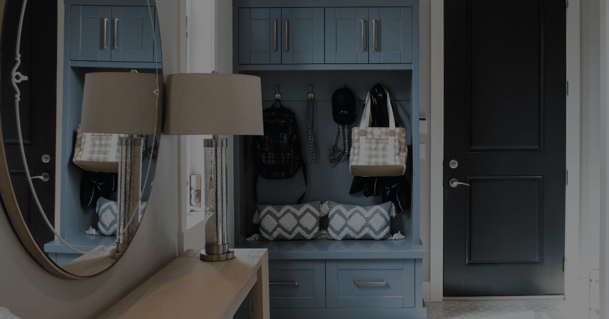 A mudroom with a bright window to the left and light blue millwork holding bags on hooks. There's a bench to sit on to remove shoes. A light wood table and mirror is to the left to catch keys and bags. A dark door is along the far wall.