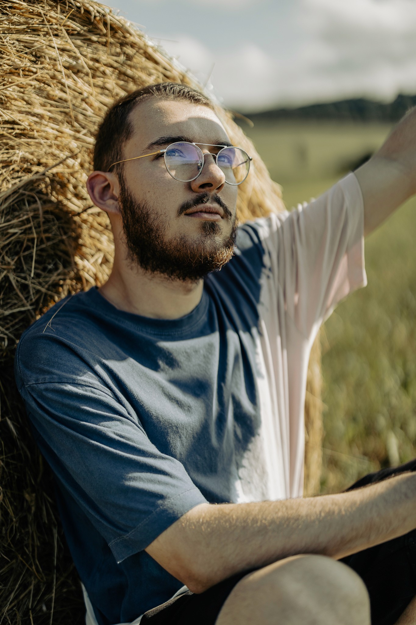 Férfi ül szalmabála mellett, napsütéses mezőn, laza nyári öltözetben.