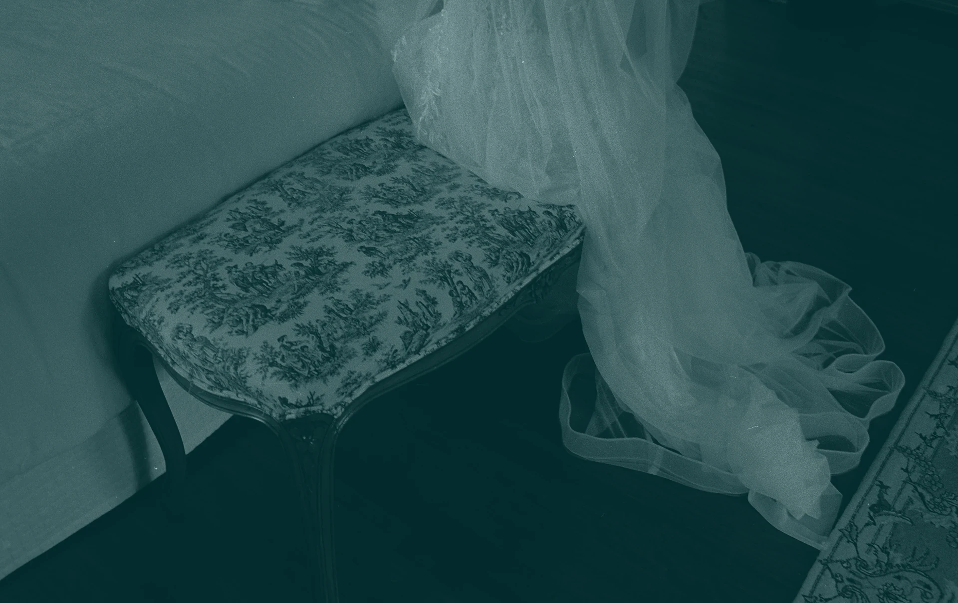 Teal-tinted photo of a bench upholstered in toile fabric, with sheer white tulle spilling over its side