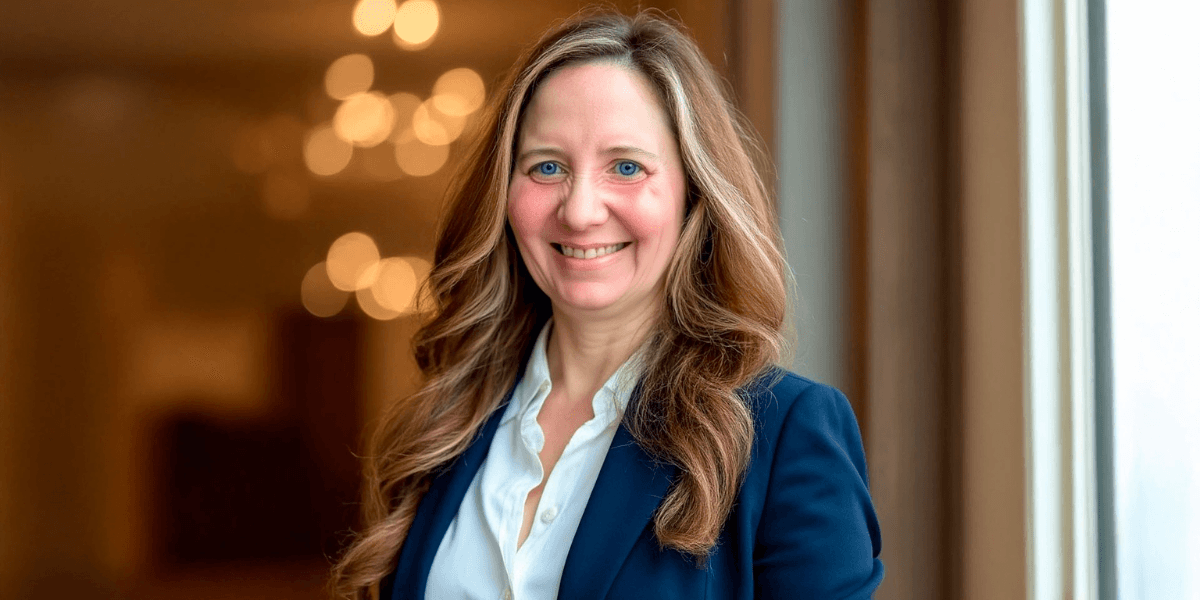 Sheera Eby, AI transformation consultant, smiling confidently in a professional blue jacket, with blurred lights in the background