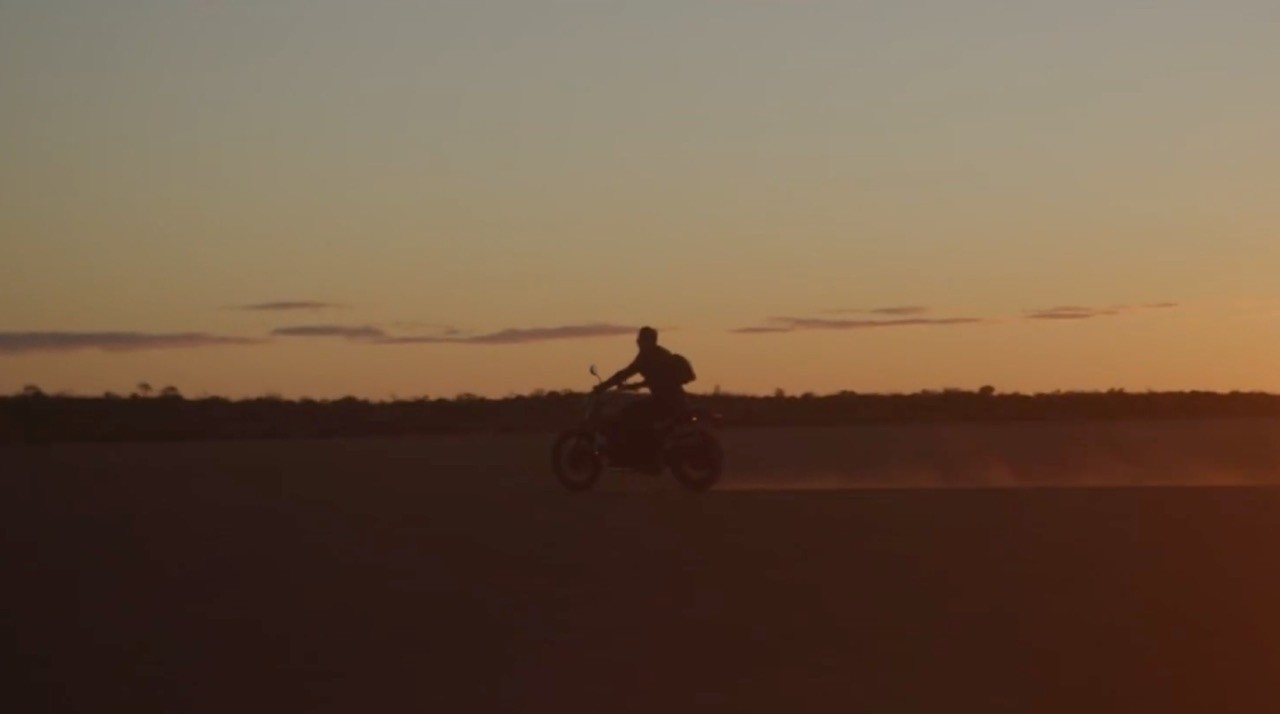Silhouette of a person on a motorcycle riding across a flat desert landscape at sunset or dusk.
