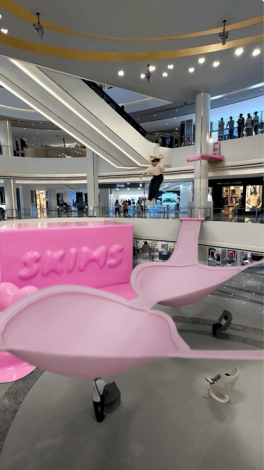 A surreal mixed-reality scene in a mall shows a person jumping onto a giant, inflatable pink SKIMS bra