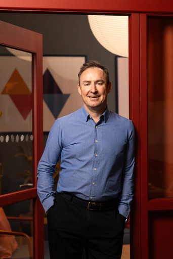 Photo of Charlie Hoult wearing a blue shirt with is hands in his pockets smiling at the smaera while leaning on an open door frame.