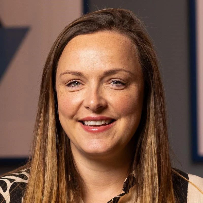 A person with long straight hair wears a patterned black-and-beige top. They are indoors with geometric wall art behind them.