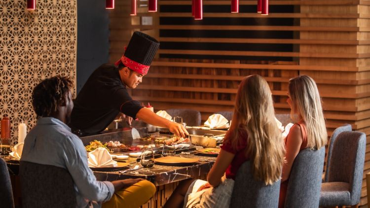 Ein Koch bereitet in einem Restaurant frische Speisen auf einem Teppanyaki-Grill direkt vor den Gästen zu, in einem modern gestalteten Ambiente mit Holzakzenten.
