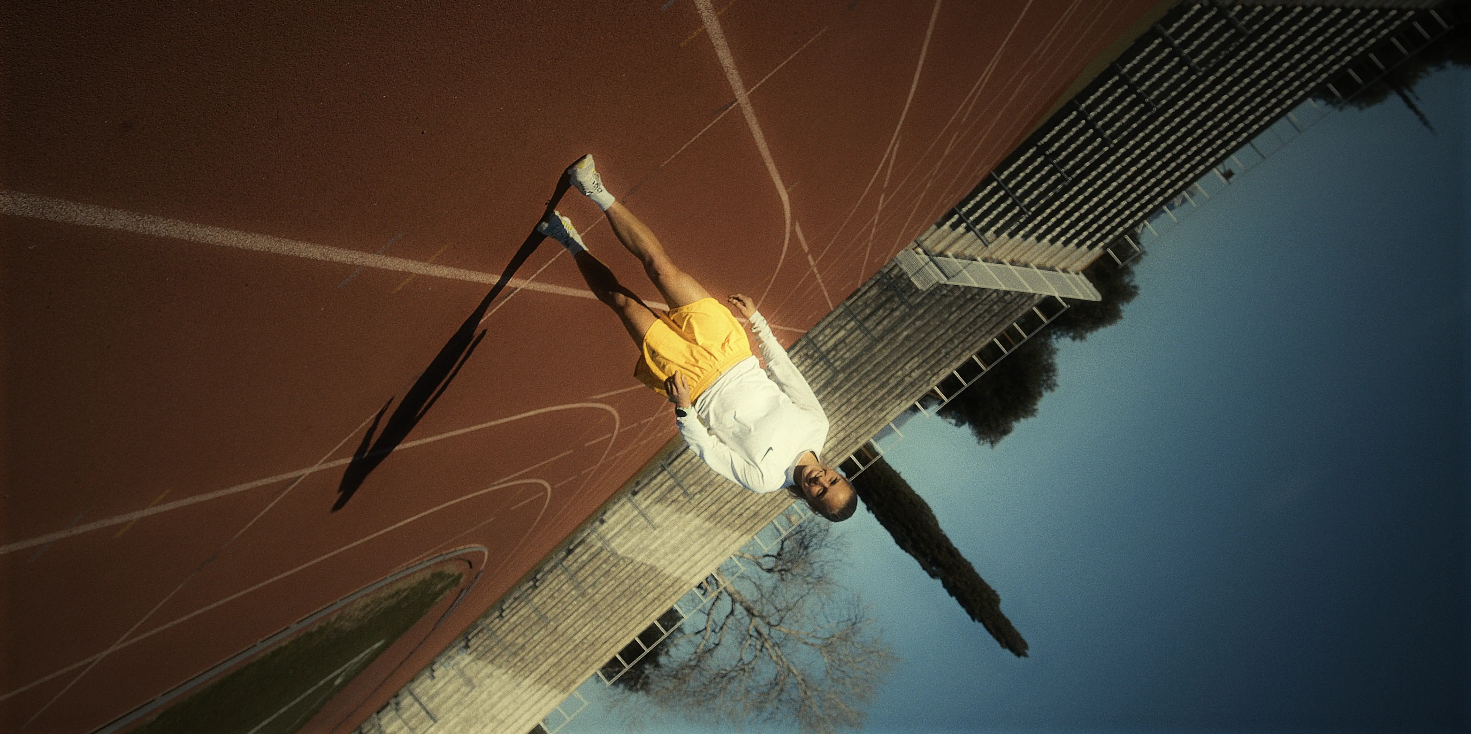 A person in a white sweater and yellow shorts walks on a sunlit outdoor track, surrounded by stadium seating under a clear blue sky.