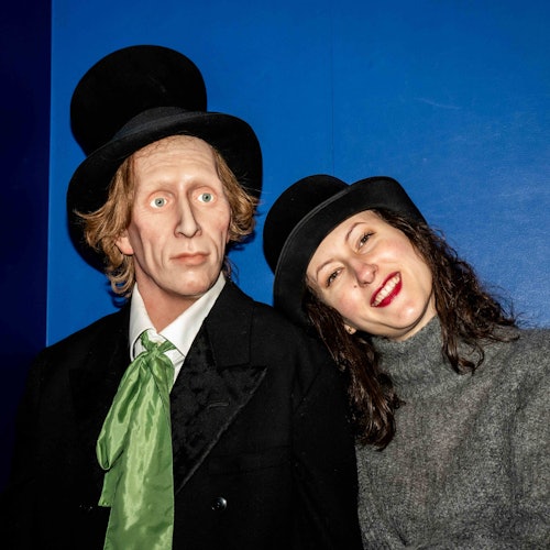 En kvinde iført en høj hat og en grå sweater smiler, mens hun læner sig op ad en voksfigur af en mand med en høj hat og et grønt slips mod en blå baggrund.