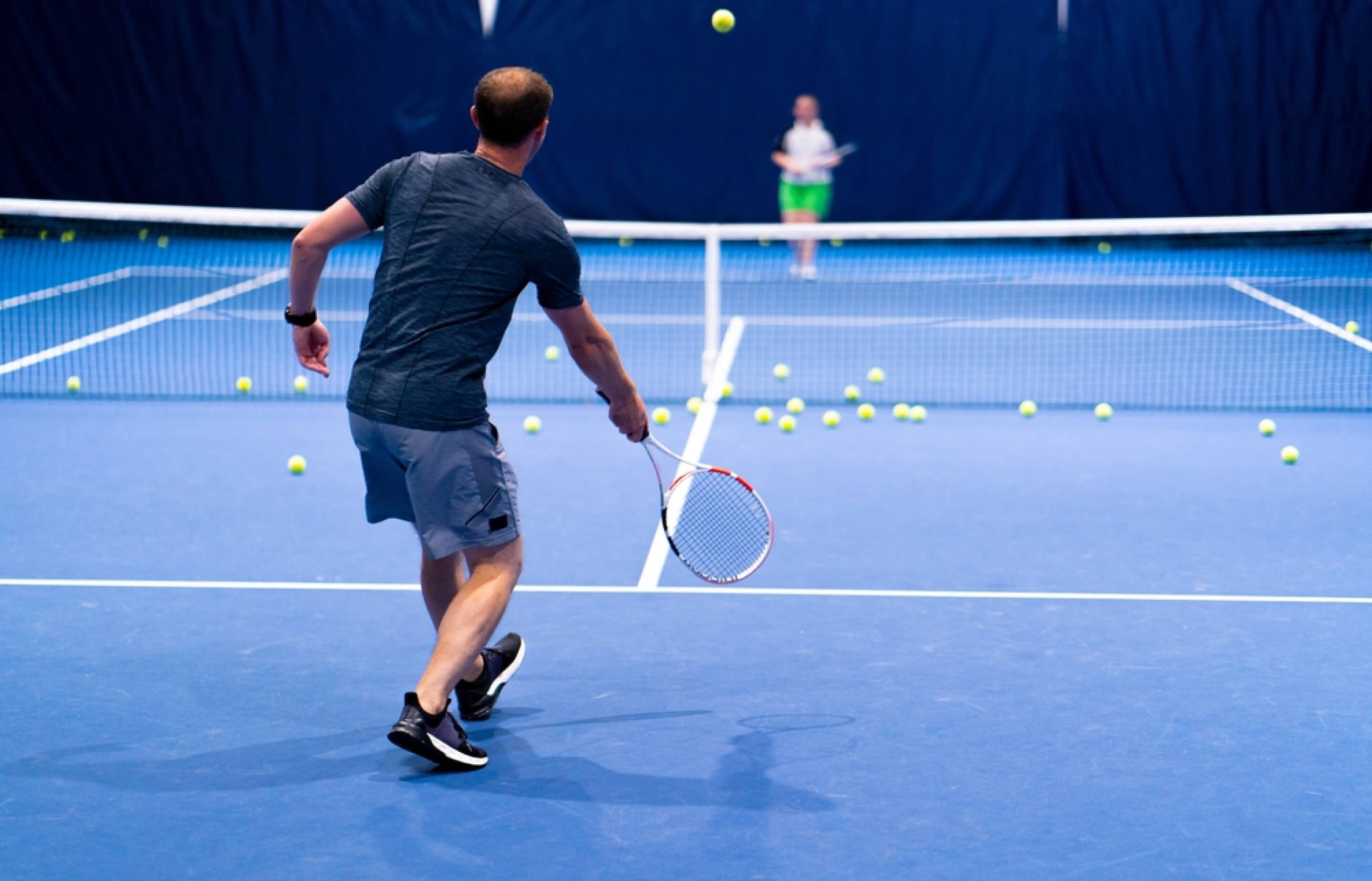 A tennis player in a dark outfit prepares to hit a ball on a blue court, with another player in the background.