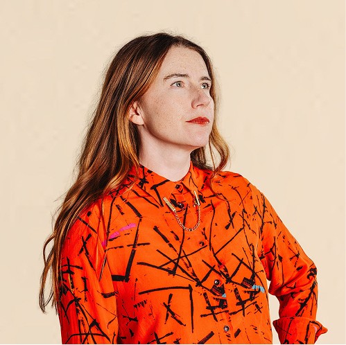 Woman with long hair wearing an orange patterned button-up shirt, looking off to the side against a neutral background.