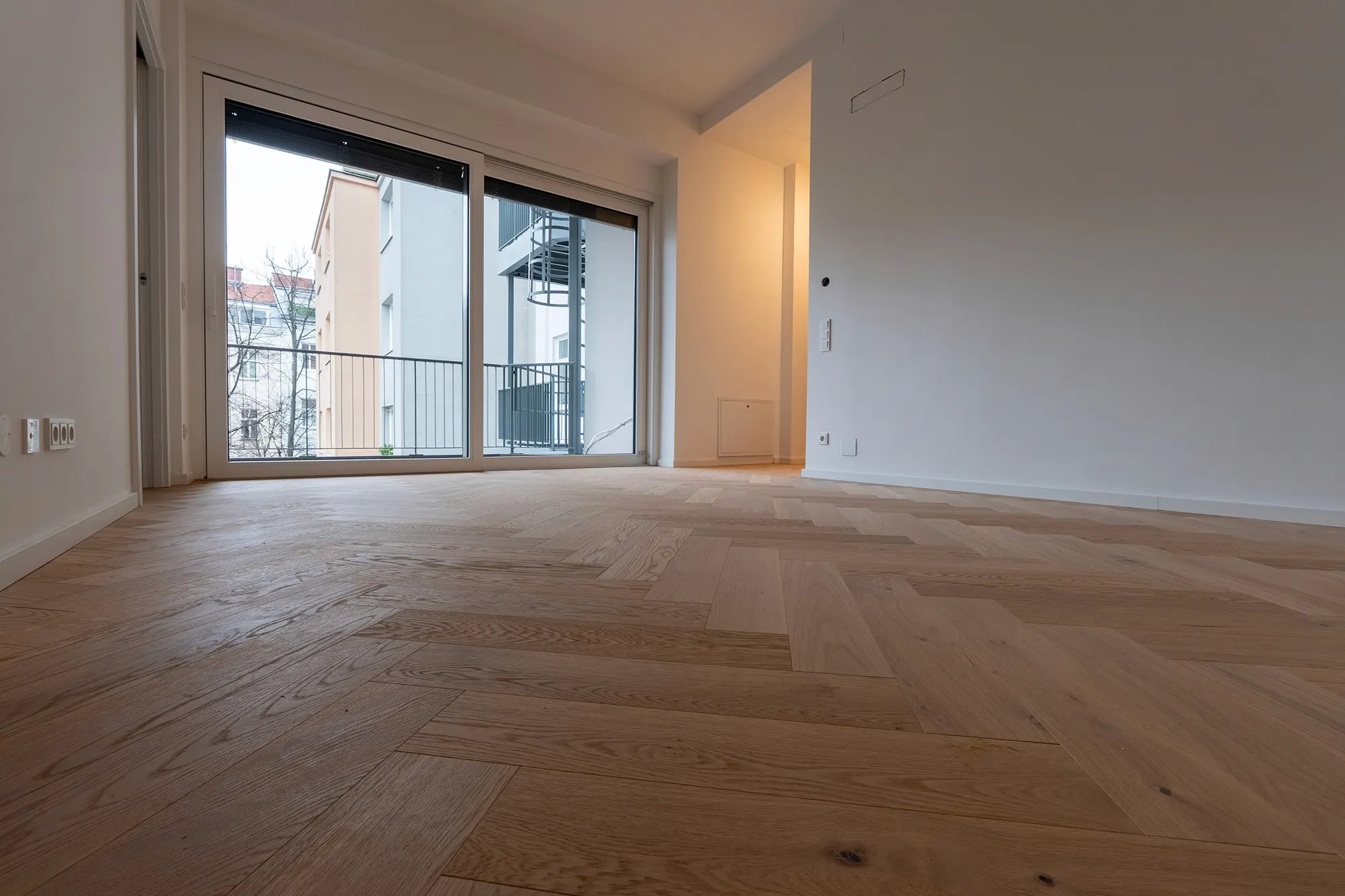 Heller Wohnraum mit Zugang zum Balkon und frisch verlegtem Fischgrät-Parkettboden.