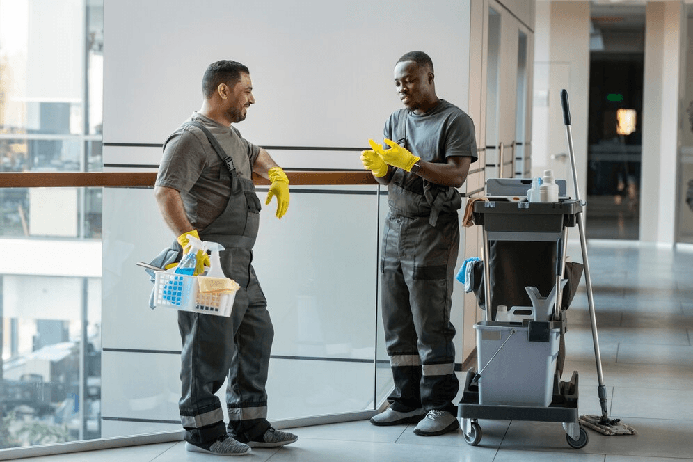 Two male janitors in uniform and yellow gloves discussing cleaning tools in a modern indoor facility with cleaning carts.