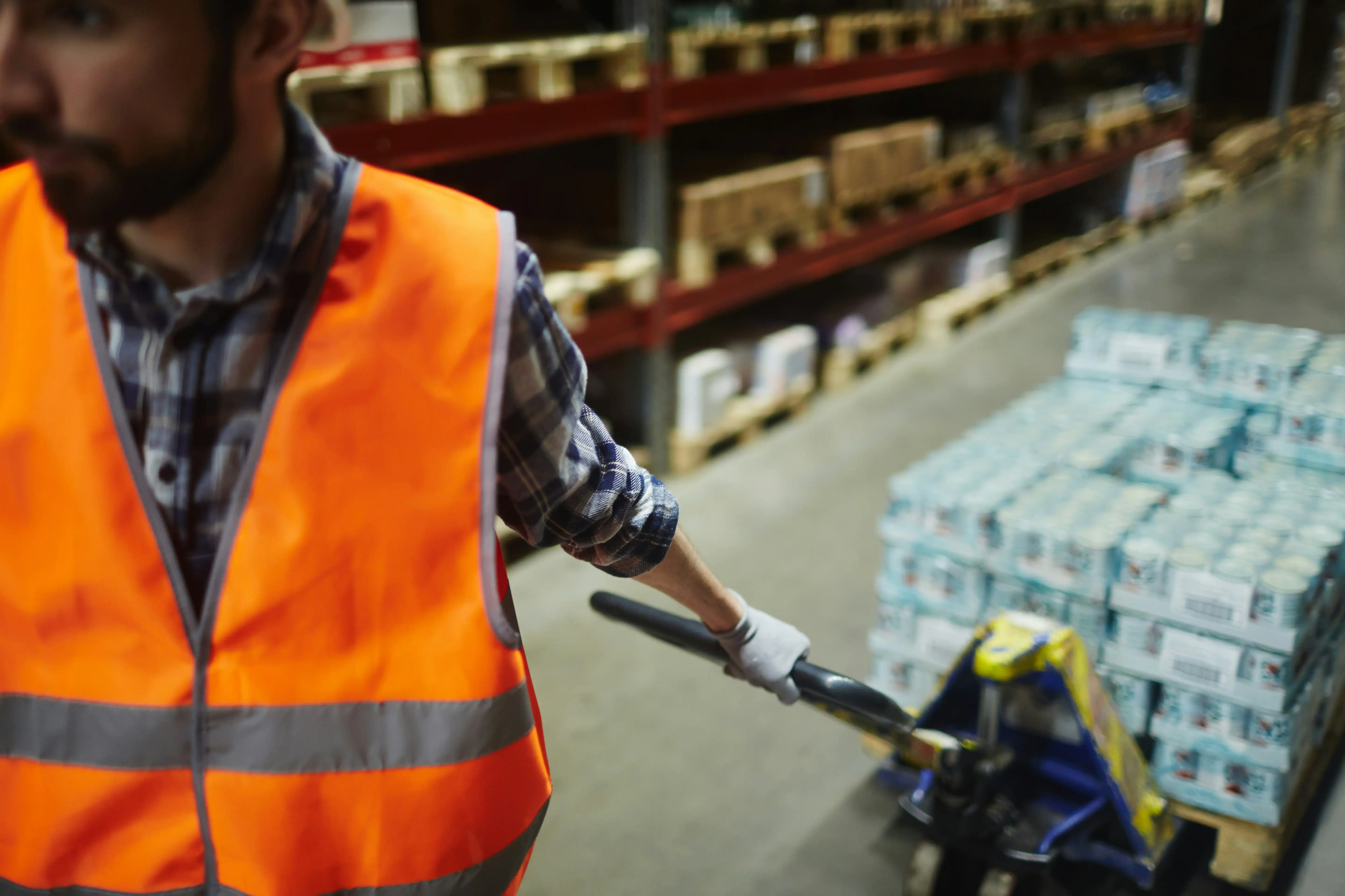 warehousre worker wearing orange high-vis jacket drags a pallet trolley behind him
