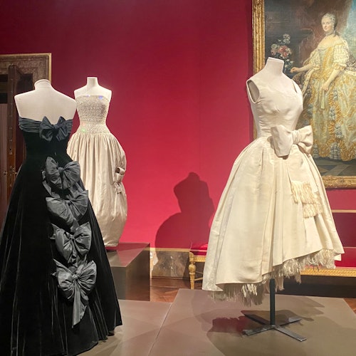 Three mannequins display elegant vintage dresses in a museum. A black dress with bows, a cream dress, and a white dress are shown.