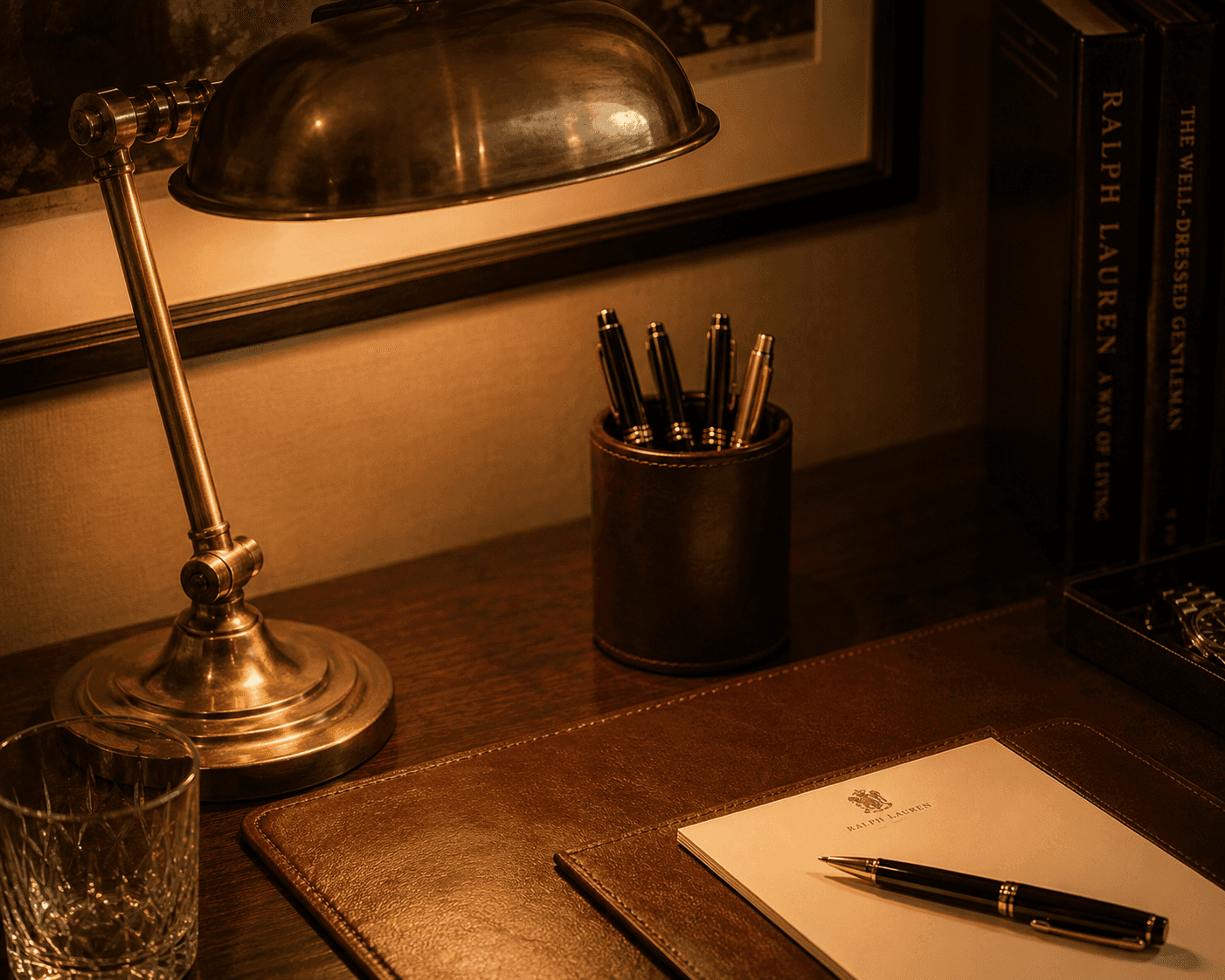 A warm, dimly lit luxury desk setup featuring a vintage brass bankers lamp illuminating a wooden desktop. A brown leather desk blotter holds a Ralph Lauren stationary notepad and a classic black and gold pen. In the background, a leather pen holder holds multiple luxury pens next to stacked books, including "Ralph Lauren: A Way of Living."